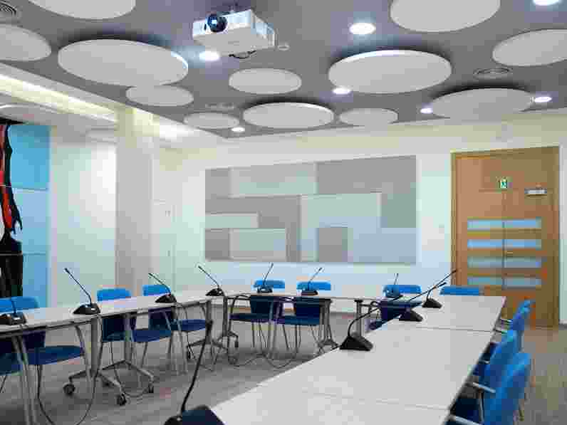 Large conference room with a U-shaped seating arrangement, grey acoustic wall panels in a pattern and white circle shaped free-hanging ceiling panels. 