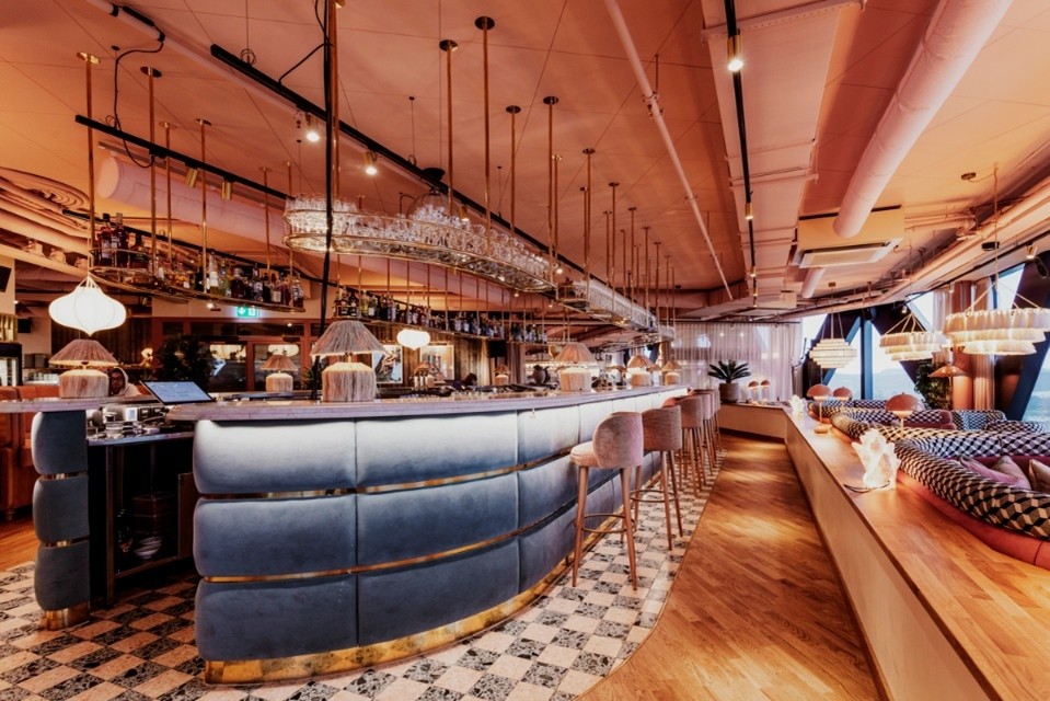 Intérieur d’un bar-restaurant contemporain avec grand comptoir courbé bleu, luminaires suspendus et ambiance chaleureuse aux tons dorés.