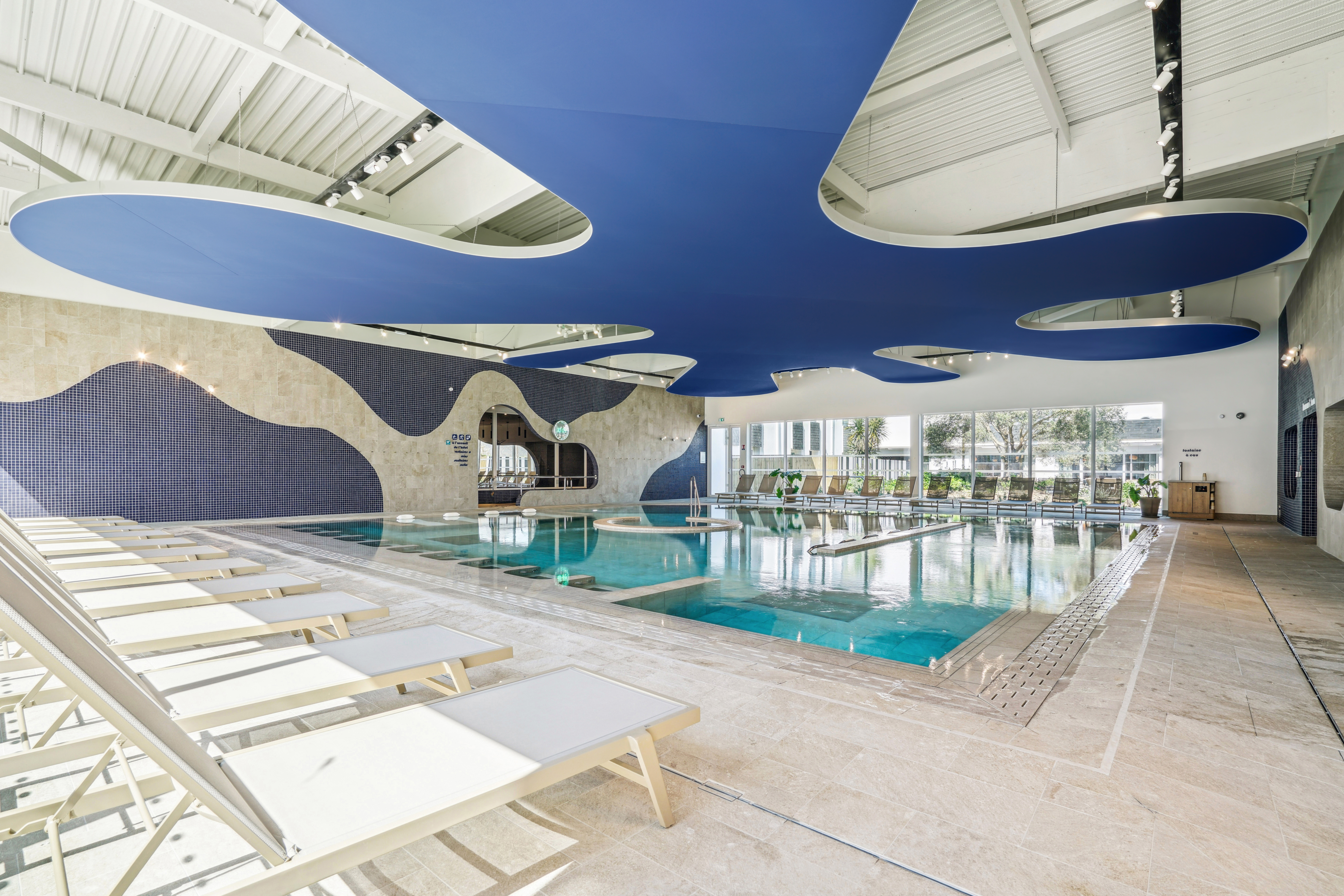 Grand espace spa lumineux du Mercure Quiberon avec piscine intérieure, plafond décoratif bleu en forme d’éclaboussure signé Ecophon Clipso™, transats clairs au premier plan et larges baies vitrées offrant une vue sur l’extérieur.