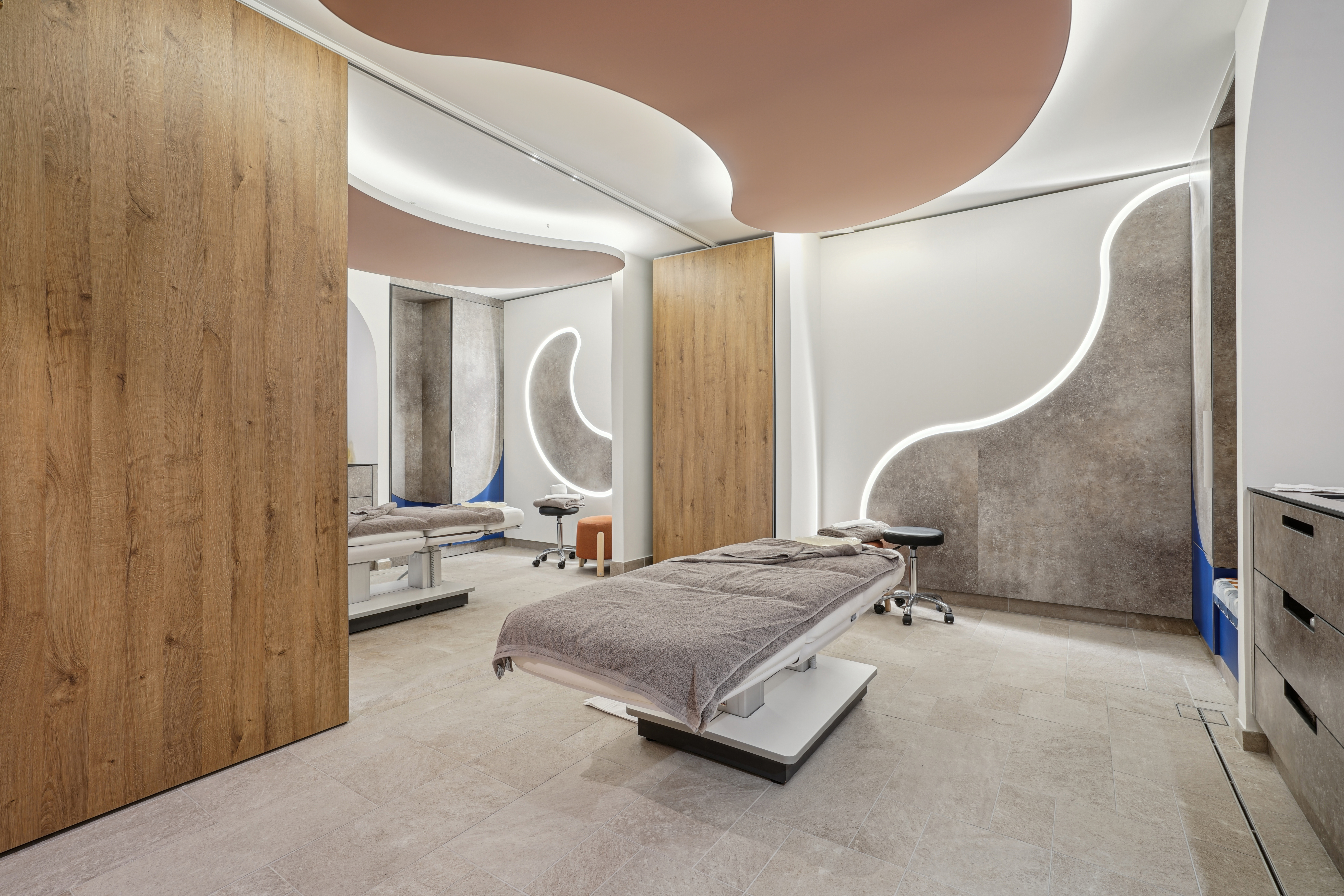 Salle de soins du spa avec deux tables de massage, murs texturés, éclairage LED courbe et éléments de plafond suspendus en forme organique pour améliorer l’acoustique.