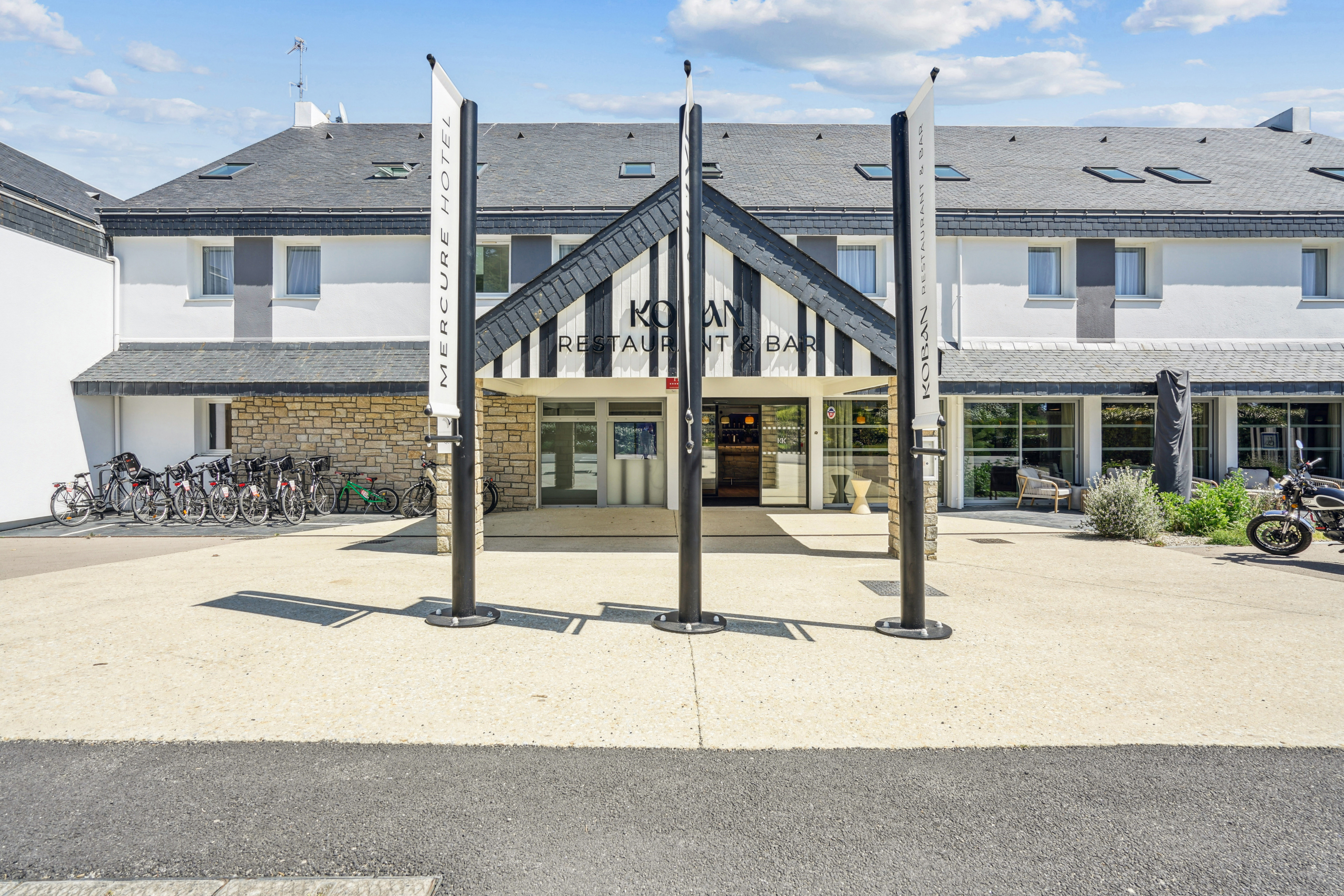 Façade du Mercure Quiberon avec entrée principale du restaurant et bar, structure en bois noir et blanc, vélos stationnés sur le côté et ciel dégagé.