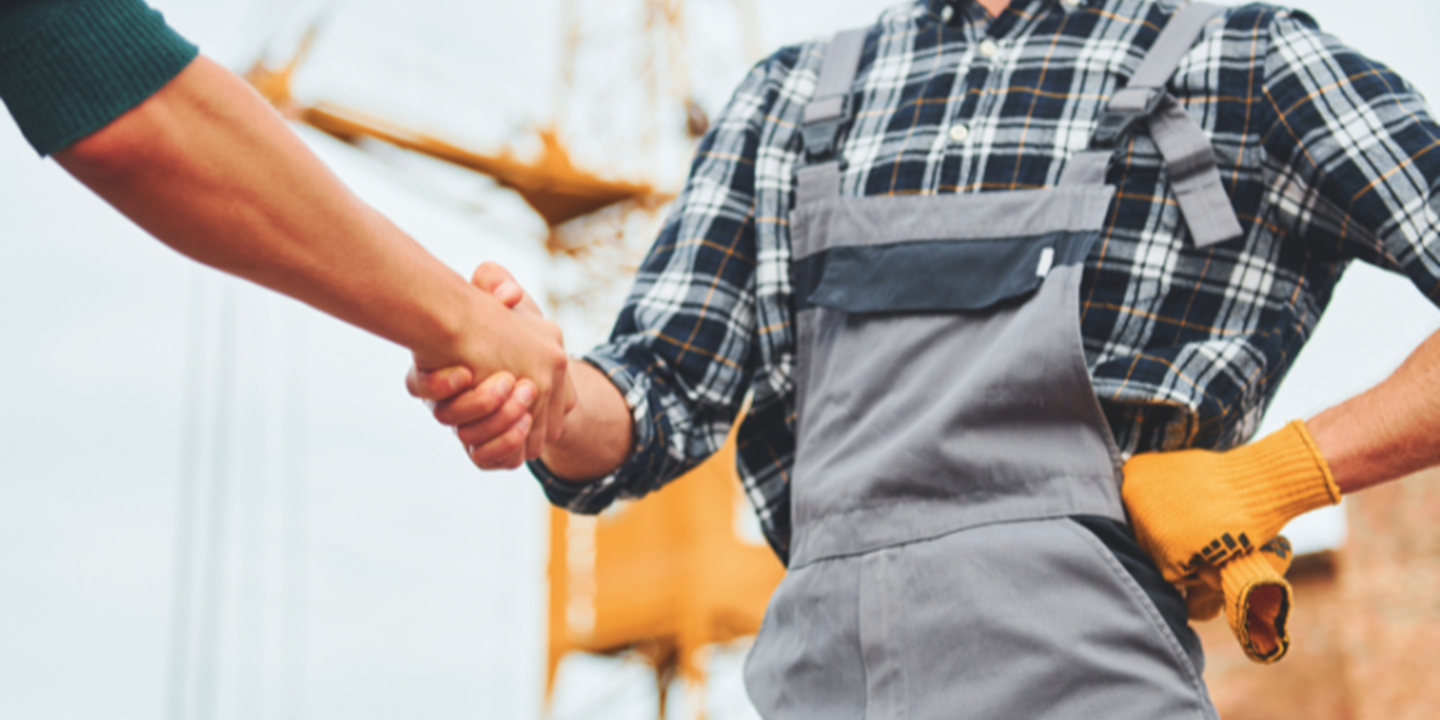 Poignée de main entre deux personnes sur un chantier, dont l’une porte une chemise à carreaux et une salopette de travail.