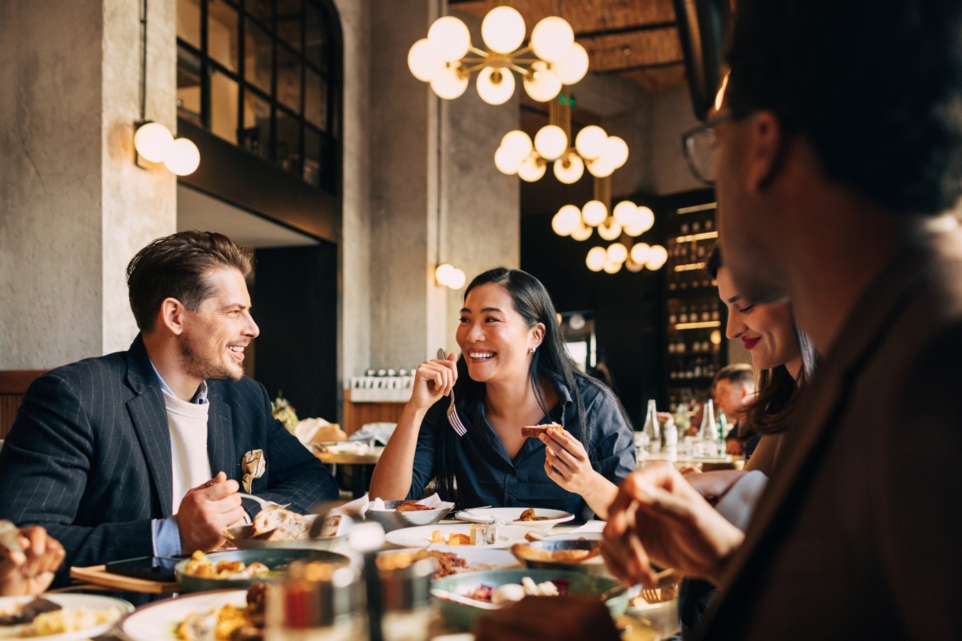 Groupe de collègues partageant un repas dans un restaurant convivial, discutant autour d’une grande table garnie de plats.