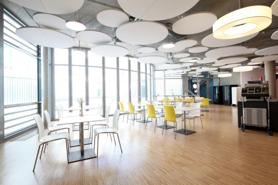 Espace de restauration lumineux avec panneaux acoustiques circulaires suspendus, mobilier jaune et blanc et grandes fenêtres.