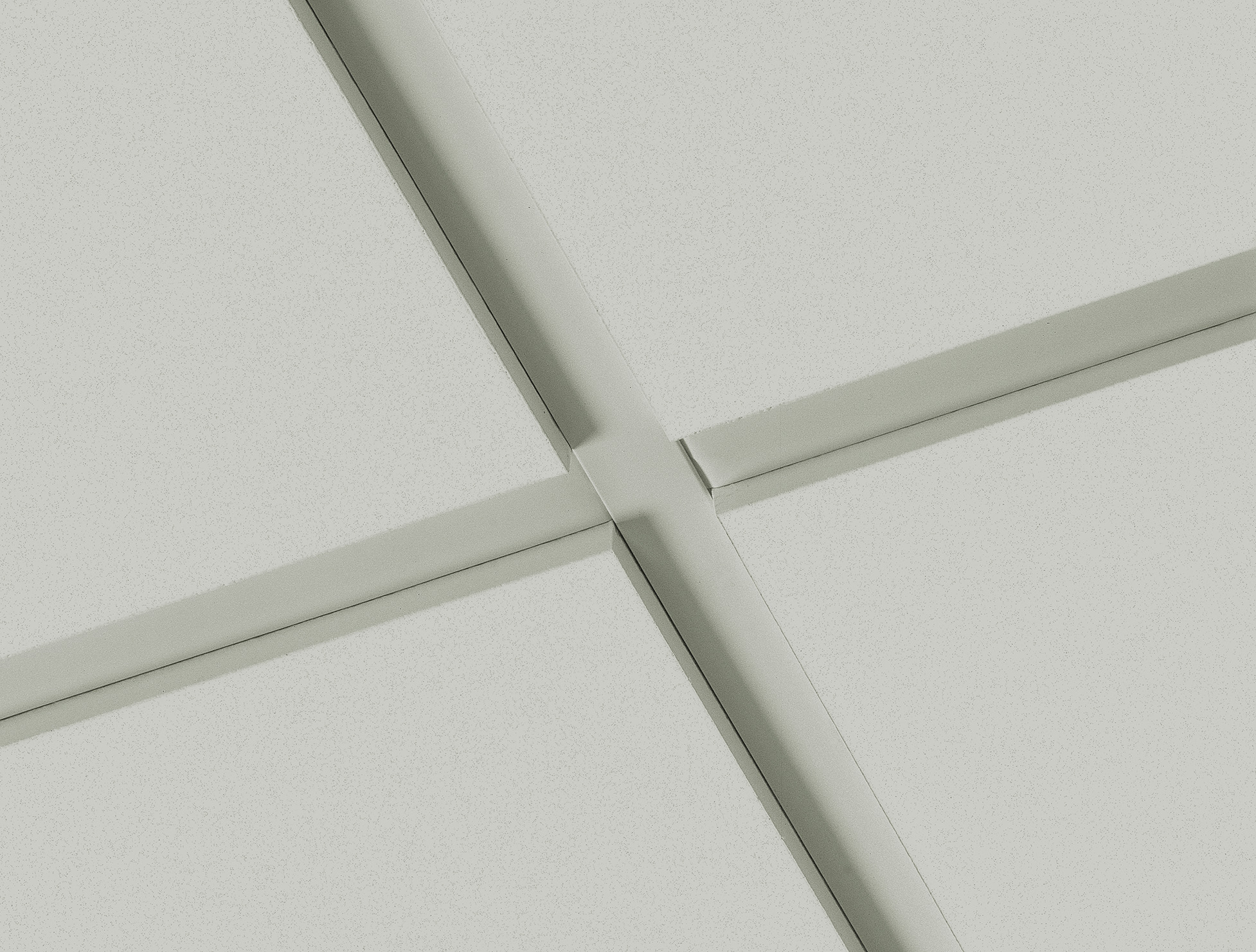Close-up view of a light grey ceiling with intersecting metal grid lines forming square panels, typical of a suspended ceiling in an office or commercial building.