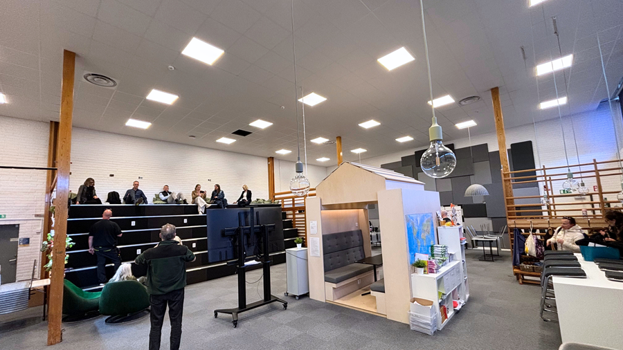 Et rummeligt, moderne rum med højt til loftet, hvor personer sidder på sorte, trappeformede bænke. Rummet indeholder en lille trækonstruktion, der ligner et hus, samt forskellige siddeområder med borde og stole. Kraftig loftsbelysning oplyser hele rummet.