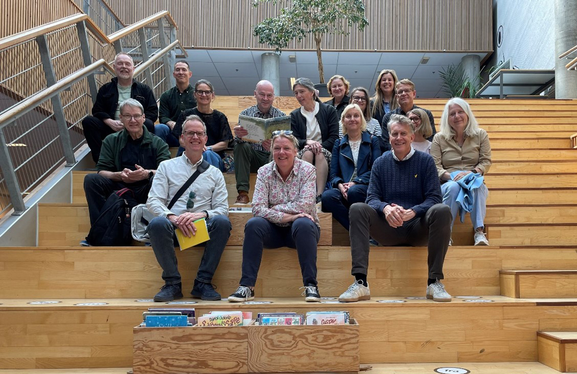 En gruppe på femten voksne sidder og poserer til et foto på brede indendørs trætrin – nogle smiler, og nogle holder bøger. Reoler med børnebøger er synlige foran dem.