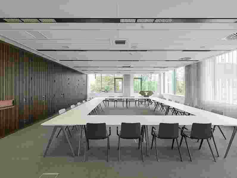 Suspended, acoustic ceiling in large meeting room with a rectangular seating arrangement, a dark wooden wall and white blinds covering part of the big windows.