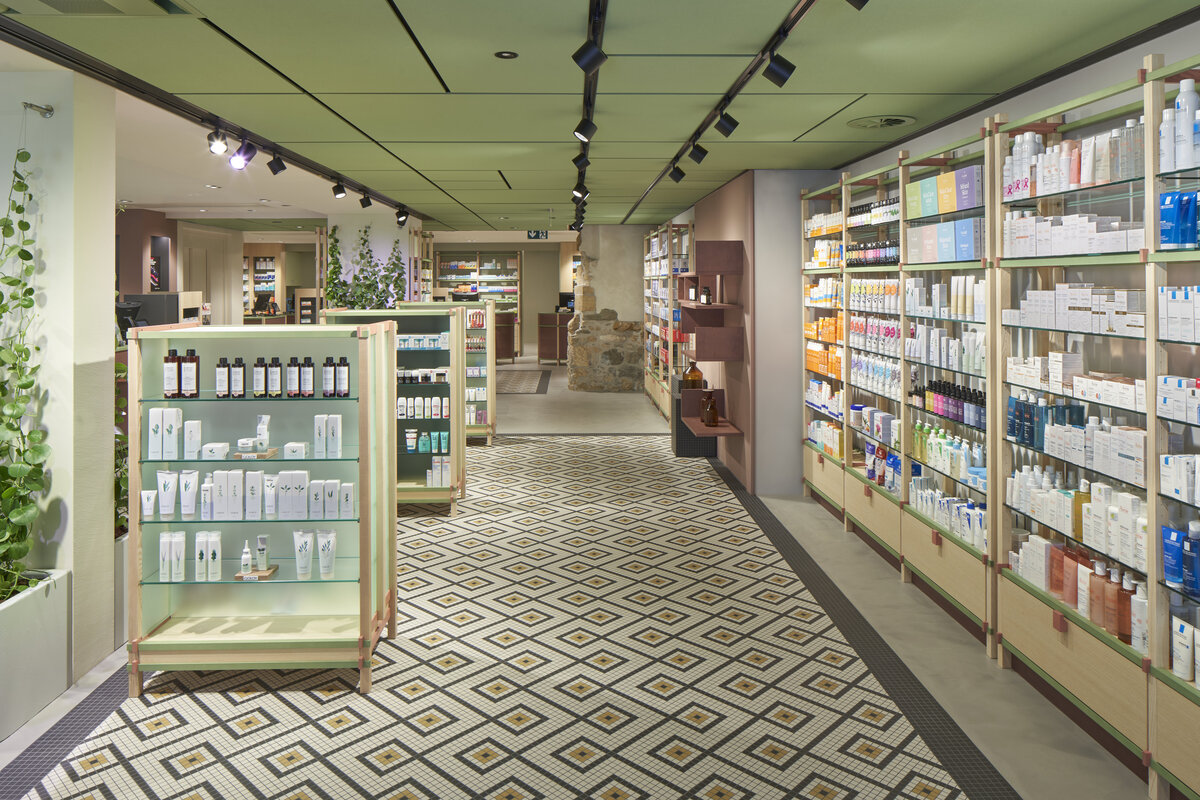 A well-lit store aisle with shelves filled with various skincare and beauty products. Green plants decorate the space, and the patterned floor has a geometric design. The green acoustic ceiling features spotlights illuminating the products.