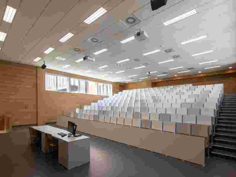White, suspended acoustic ceiling in lecture hall with wooden walls and a slanting floor with rows of chairs