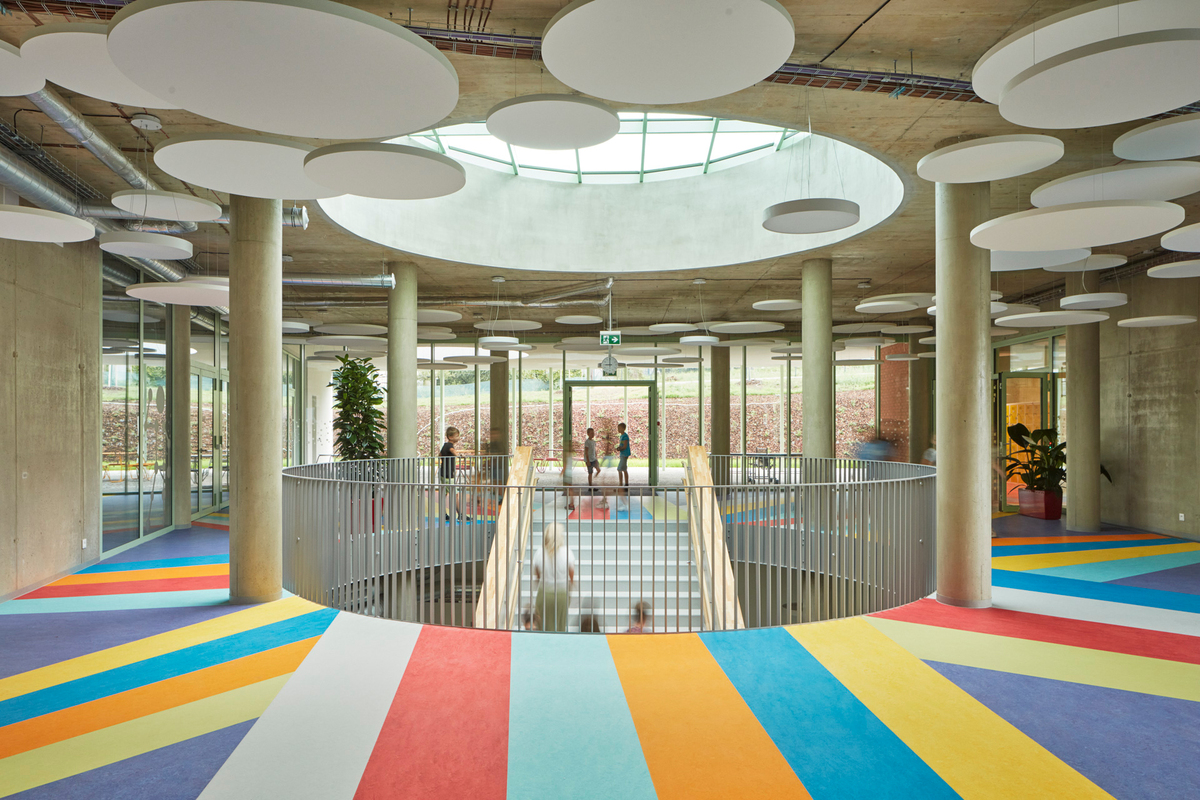 A bright, modern interior with circular ceiling panels, a central skylight, colourful striped flooring, and a round staircase surrounded by glass walls and columns. People are visible in the background.