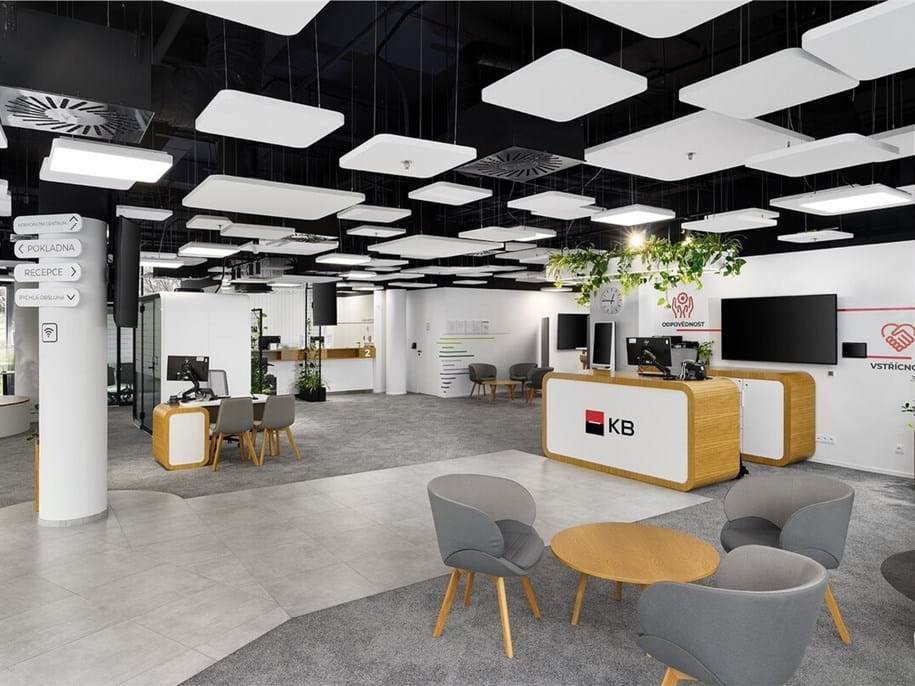 Modern open office space with reception desk, round tables and grey chairs, computer workstations, large screens, and white ceiling panels. Green plants add a touch of nature to the clean, bright interior.