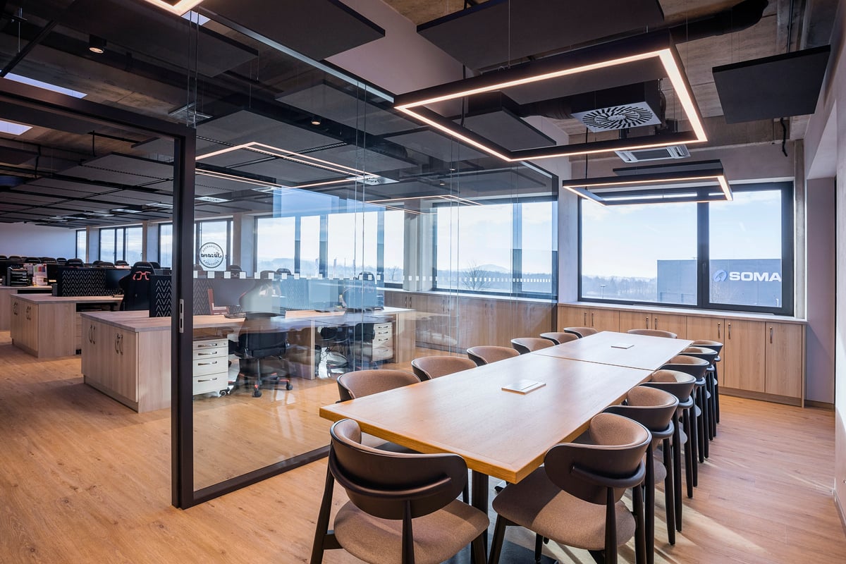 Modern office with glass-walled meeting room, long wooden table, black chairs, and open workspace beyond. Large windows let in natural light, creating a bright, airy atmosphere with sleek, minimalist design elements.