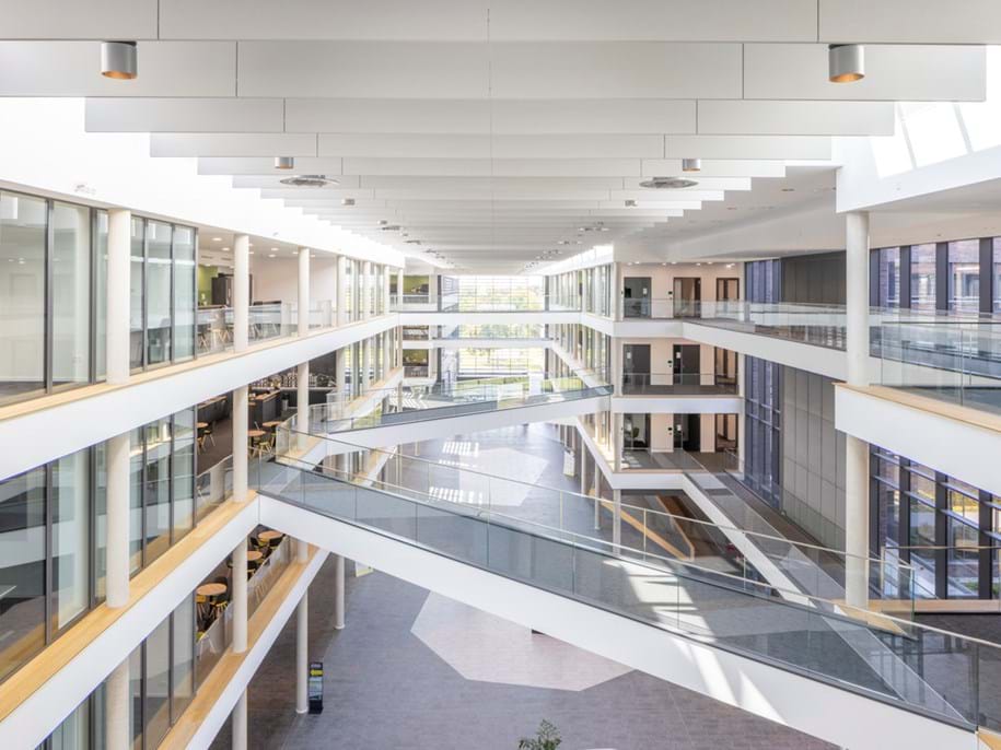 Bright, modern multi-storey building interior with large glass windows, open corridors, and multiple intersecting walkways, allowing natural light to fill the spacious atrium.