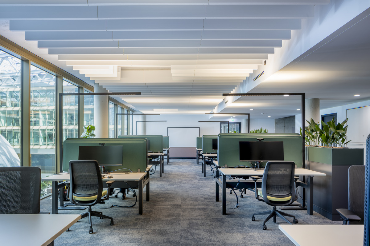 Modern open-plan office with rows of green desk partitions, ergonomic chairs, computer monitors, large windows on the left, and ceiling panels overhead. Potted plants add greenery to the bright, spacious workspace.