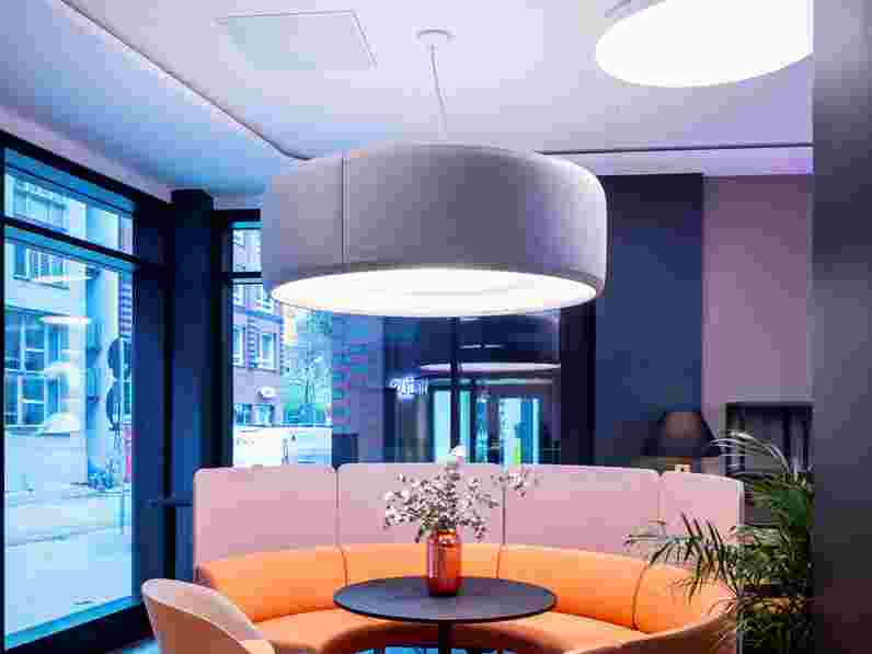 White, suspended acoustic ceiling, a swirling orange sofa and big, grey ceiling lamp in office furnture showroom 
