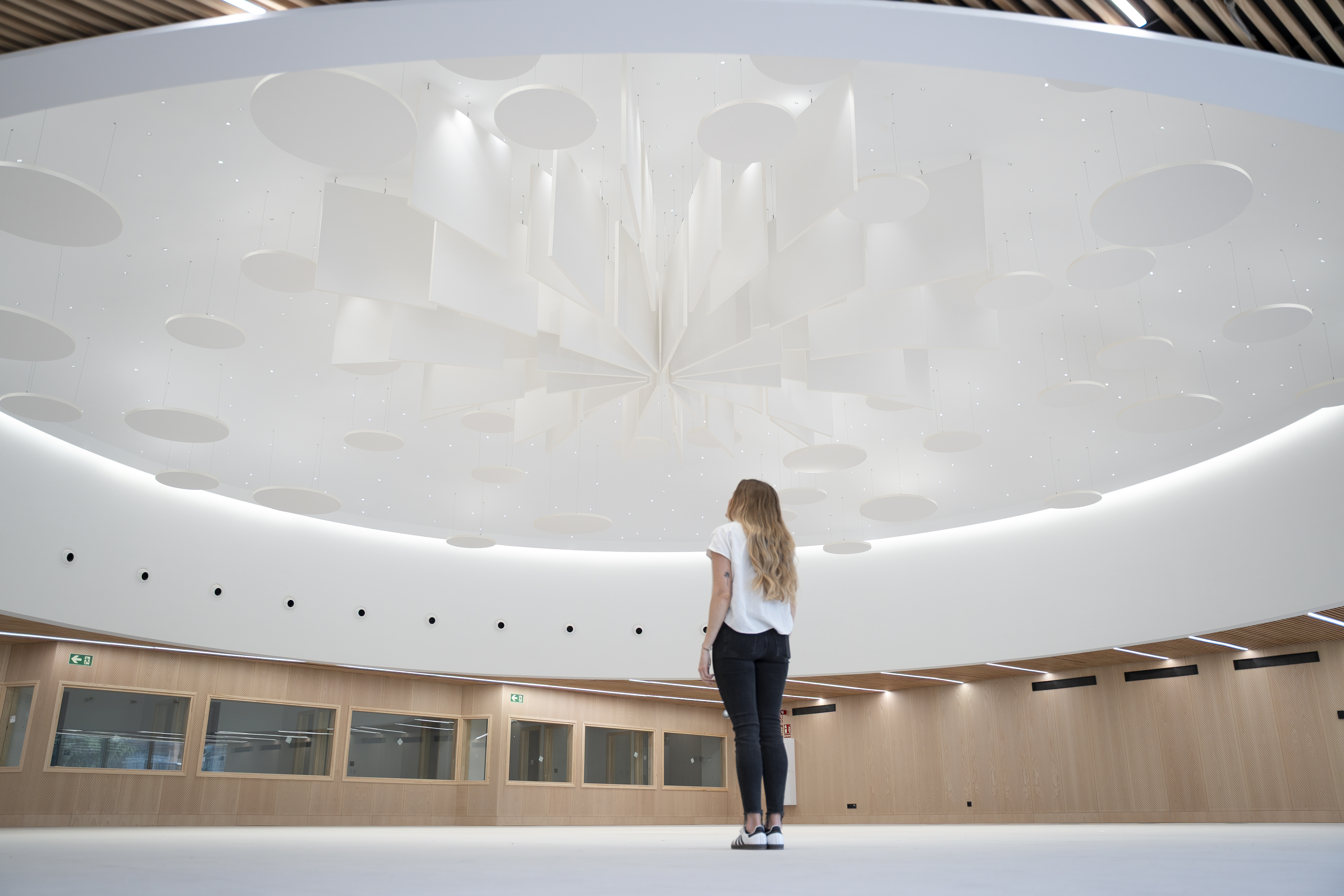 A woman with long hair stands in a modern, spacious room, looking up at a unique, white geometric ceiling installation. The room has wooden walls, large windows, and bright lighting.