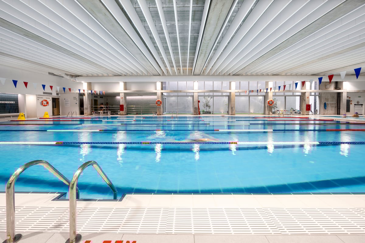 Indoor swimming pool with clear blue water, lane dividers, a metal ladder in the foreground, safety signs, and life buoys on the walls. The ceiling has white panels and large windows let in natural light.
