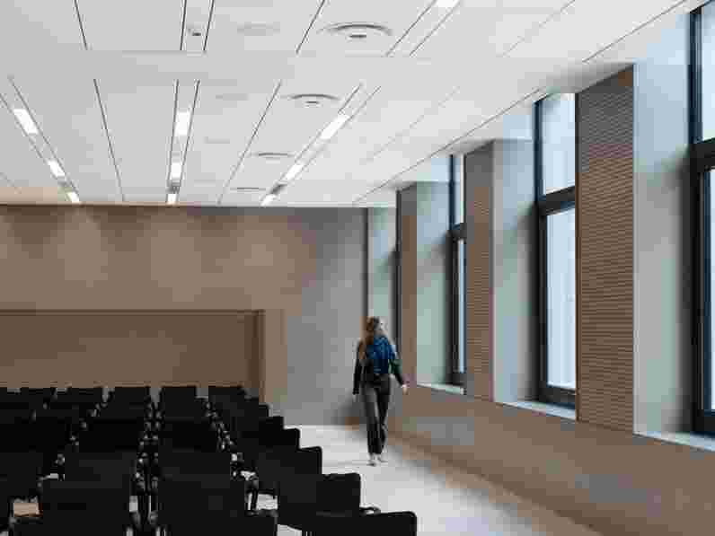 White, suspended acoustic ceiling in large conference room with beige floor and walls,  black chairs in multiple rows and a woman looking out the big windows