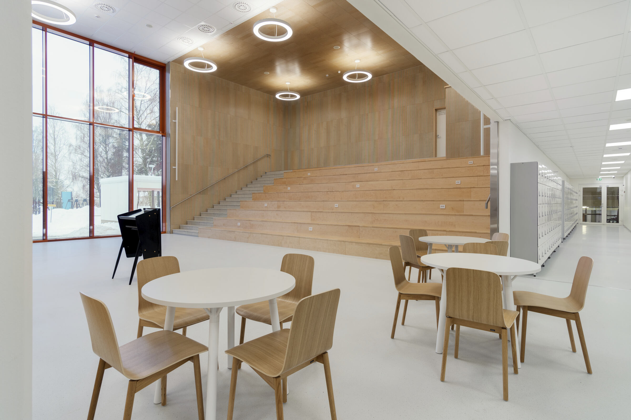 Hämeenlinnan yhteiskoulun uudisrakennuksen aula. Etualalla tuoli- ja pöytäryhmiä, taustalla puiset jättiläisen portaat. Alakatot Ecophonin valkoisia CarbonLow-akustiikkalevyjä.