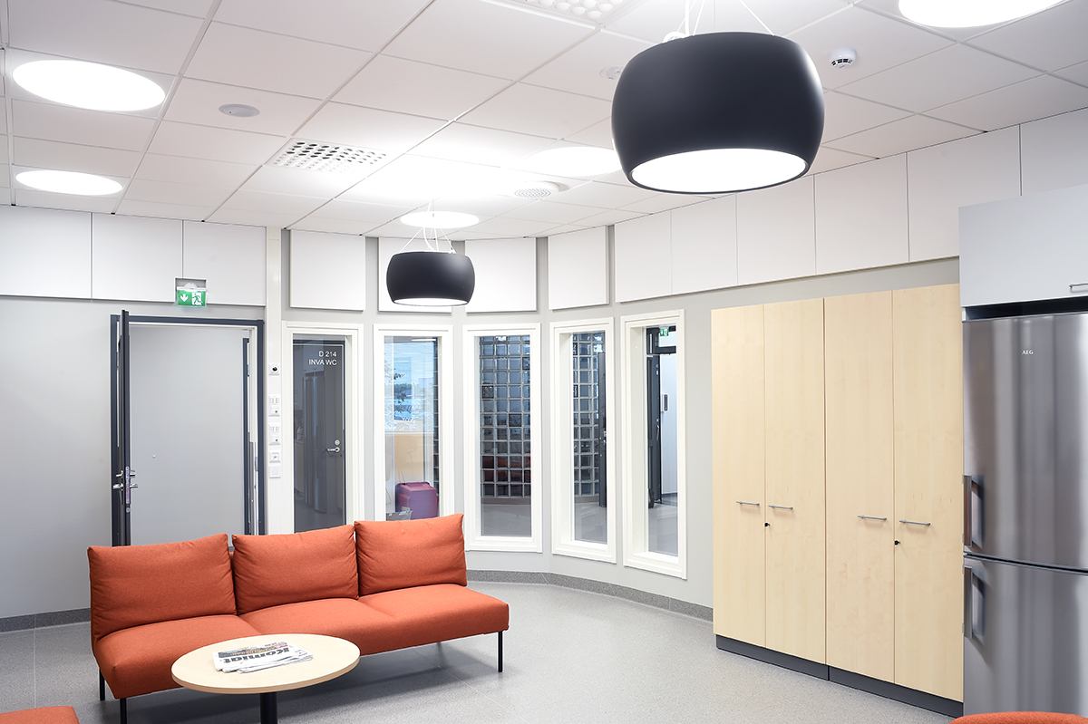 A modern lounge with orange sofas, a round table with magazines, large black pendant lights, light wood cupboards, a stainless steel fridge, and windows along a curved wall. The space is clean and brightly lit.