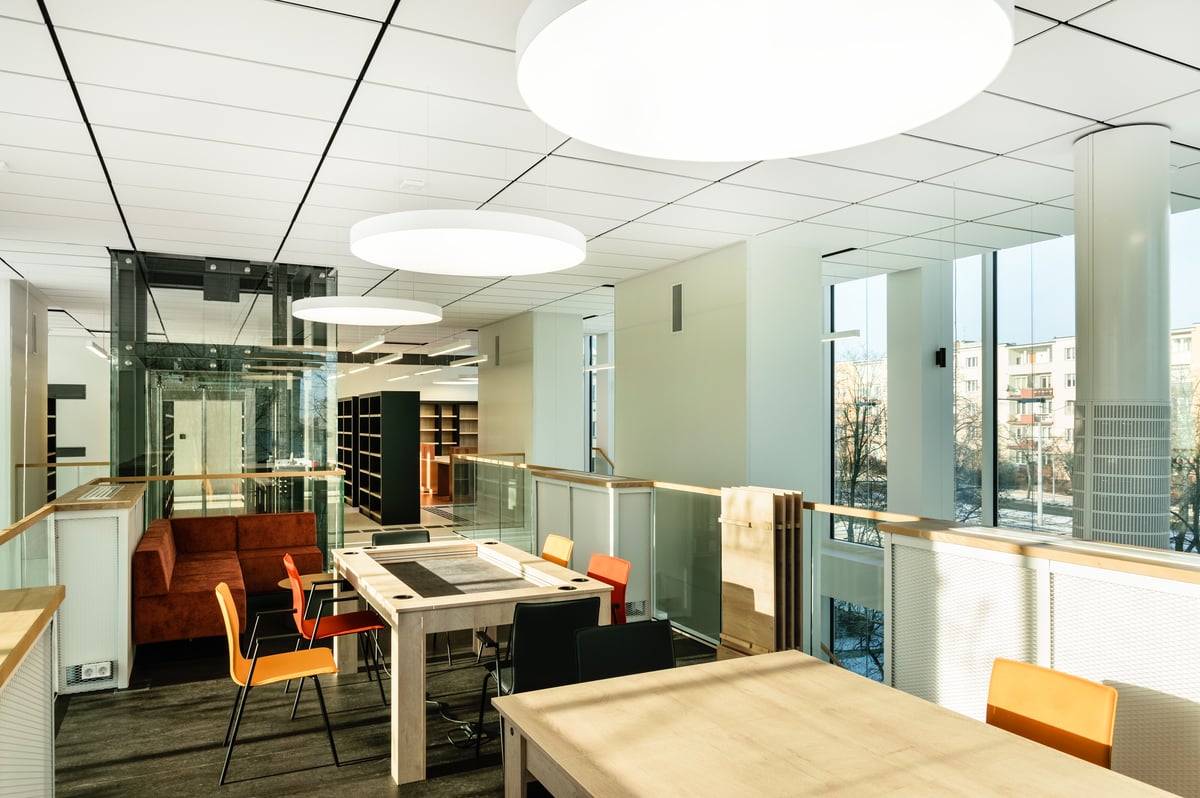 A modern, bright office with large windows, wooden tables, colourful chairs, and contemporary lighting. There are bookshelves and a red sofa in the background, with natural light streaming in.