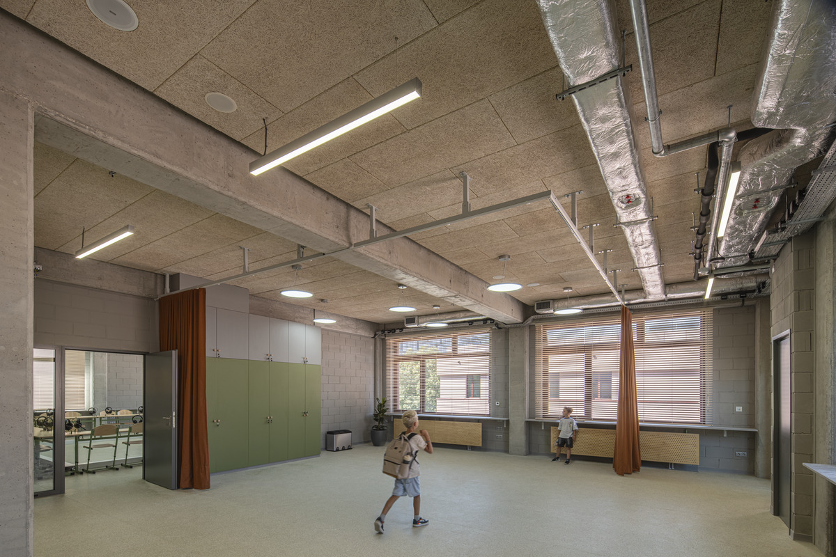 A spacious, modern classroom with exposed ceilings, large windows, green cupboards, and two children wearing rucksacks walking across the room. The space has natural light and visible ductwork.