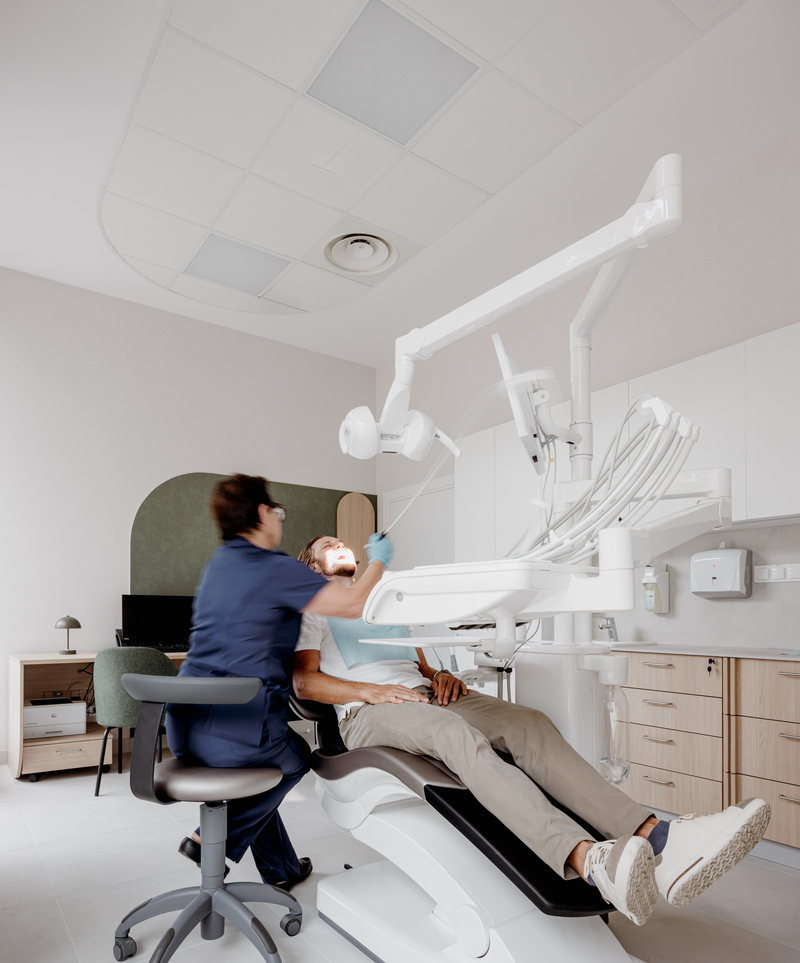 A dentist in blue scrubs examines a patient's mouth in a modern, well-lit dental surgery. The patient is reclined in a dental chair, surrounded by dental equipment and cabinetry.