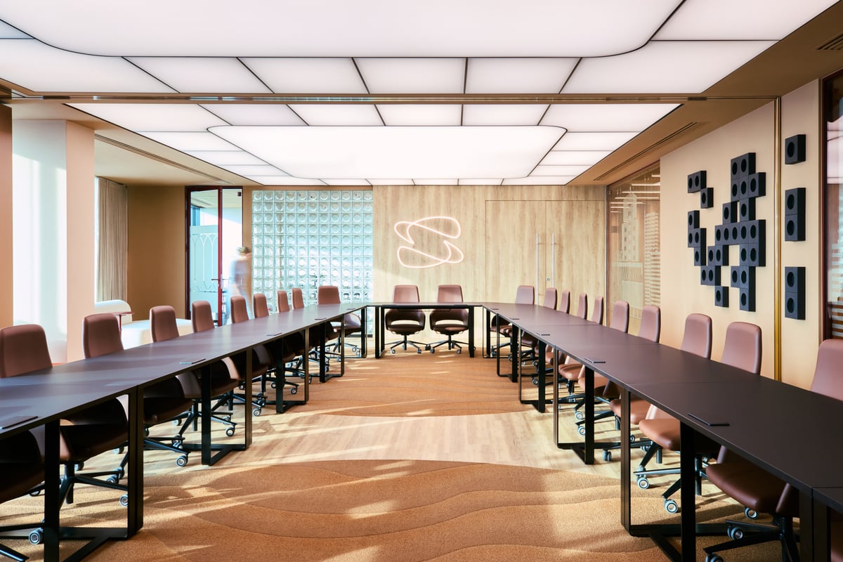 A modern conference room with two long black tables arranged in a U-shape, pink office chairs, a patterned carpet, wall art, and a glowing abstract light fitting on a wooden wall.