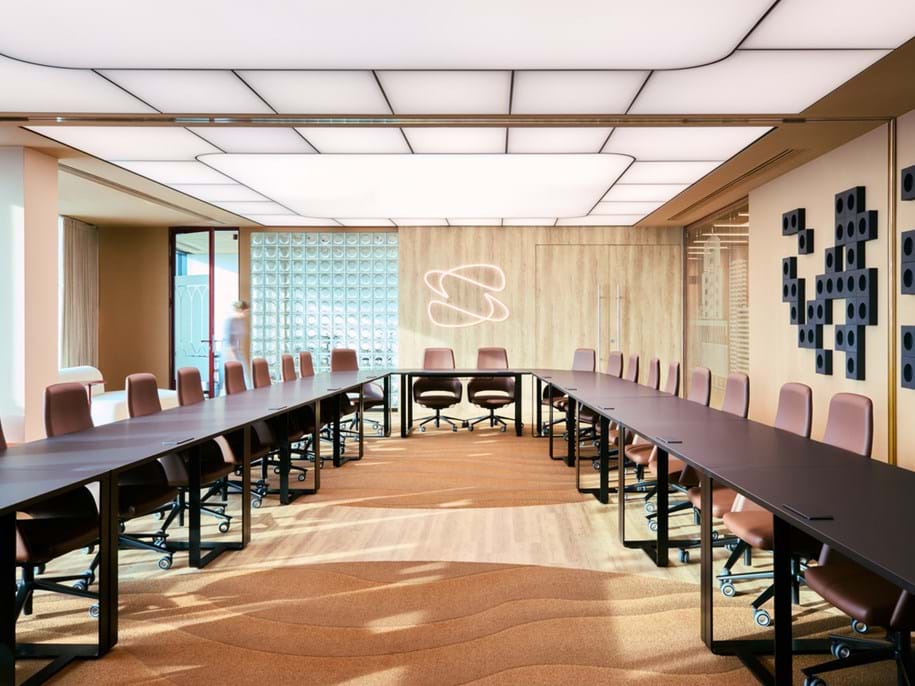 A modern conference room with two long black tables arranged in a U-shape, pink office chairs, a patterned carpet, wall art, and a glowing abstract light fitting on a wooden wall.