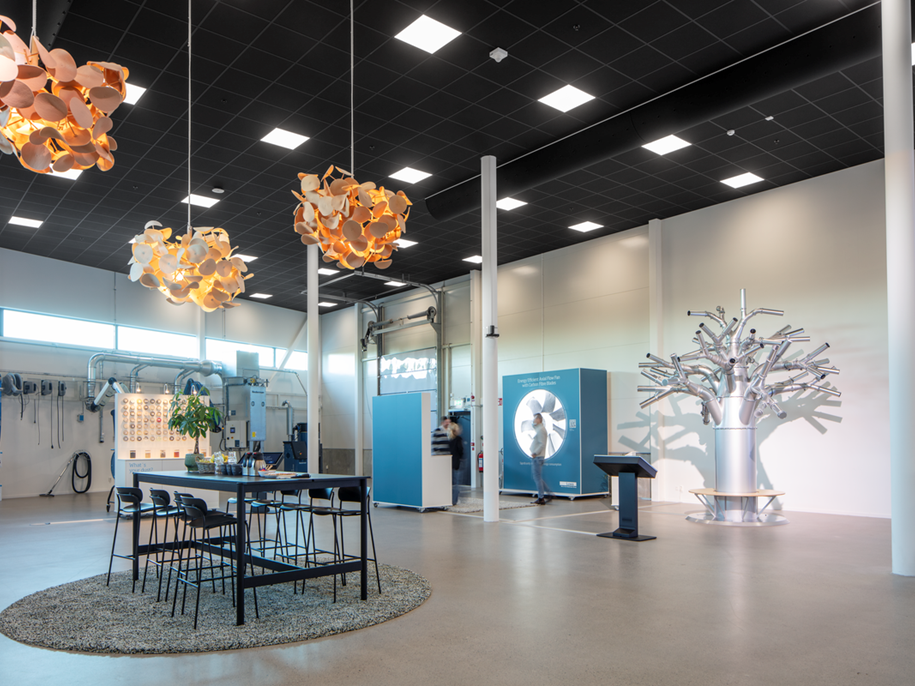 A modern, spacious exhibition room with high ceilings, hanging abstract orange lights, a table with chairs on a round rug, interactive displays, a metallic tree sculpture, and various tools mounted on the back wall.