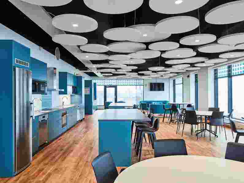 Round, free-hanging acoustic ceiling panels hanging at different levels in office kitchen and dining area with furniture and kitchen in turquoise