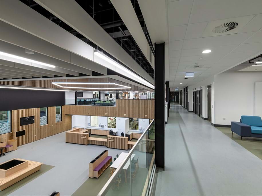Modern, bright interior space with open seating areas, wood accents, large windows, and a high ceiling. A corridor with doors and blue seating runs alongside the open atrium, separated by a glass balustrade.
