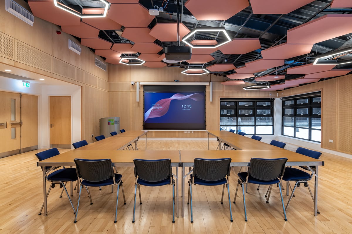 A modern conference room with two rows of tables arranged in a U-shape, black chairs, a large screen displaying 12:15, wooden floors, and hexagonal ceiling panels with geometric lights.