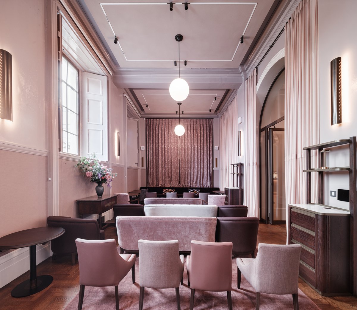 A stylish, elegant lounge with pastel pink and mauve seating, wooden floors, tall windows, soft lighting, and modern décor. A floral arrangement sits on a side table, and curtains frame an arched doorway at the back.