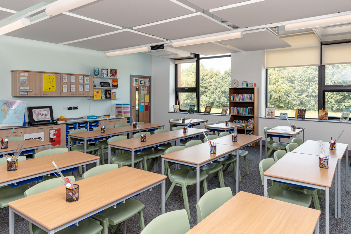 A bright, tidy classroom with several desks and green chairs, each desk holding a pencil pot. There are bookcases, wall displays, windows with views of trees, and educational materials around the room.