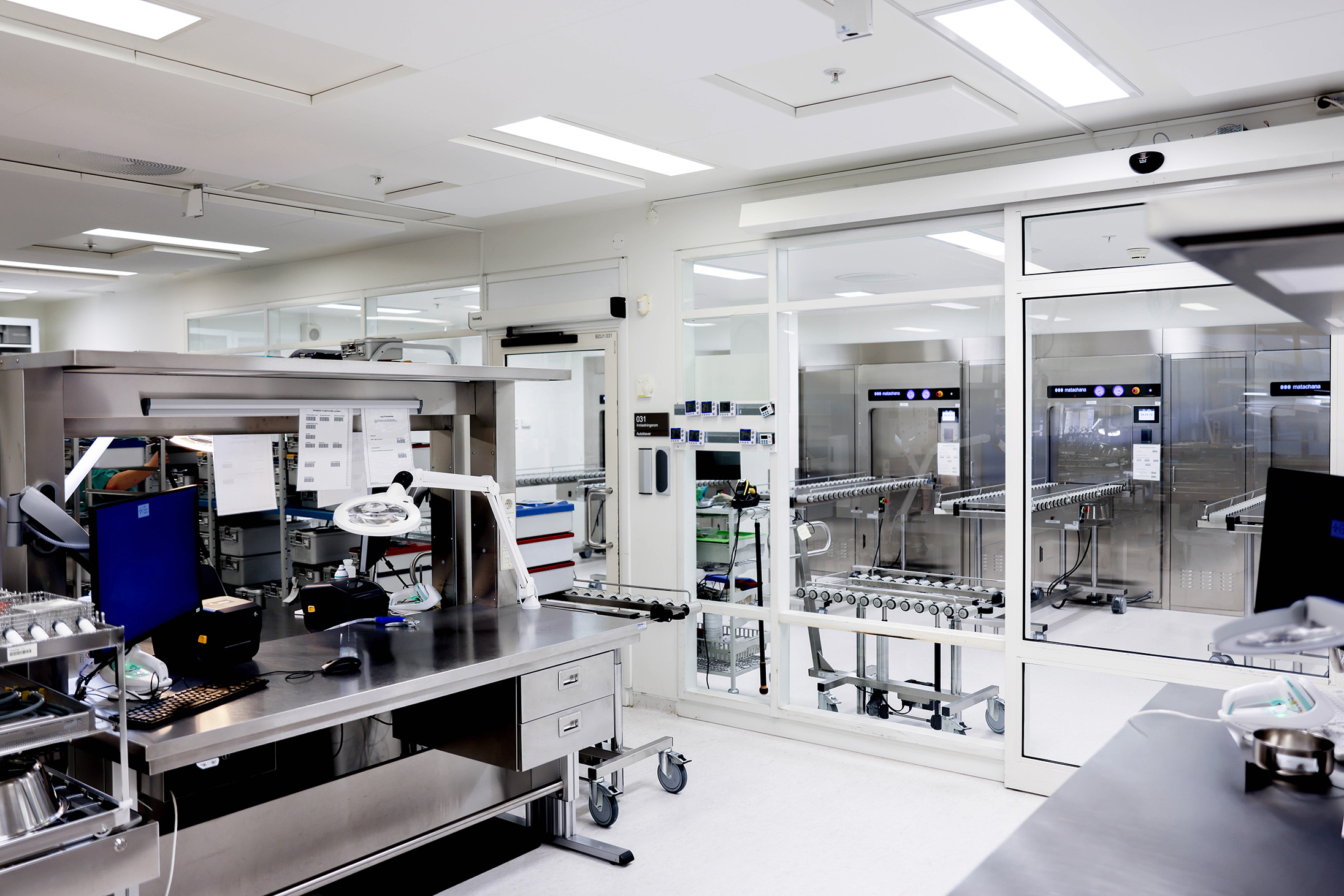 A modern, clean laboratory with stainless steel workstations, computers, and scientific equipment. Large glass windows separate the lab from another room with industrial machines and conveyor belts. Bright overhead lighting.