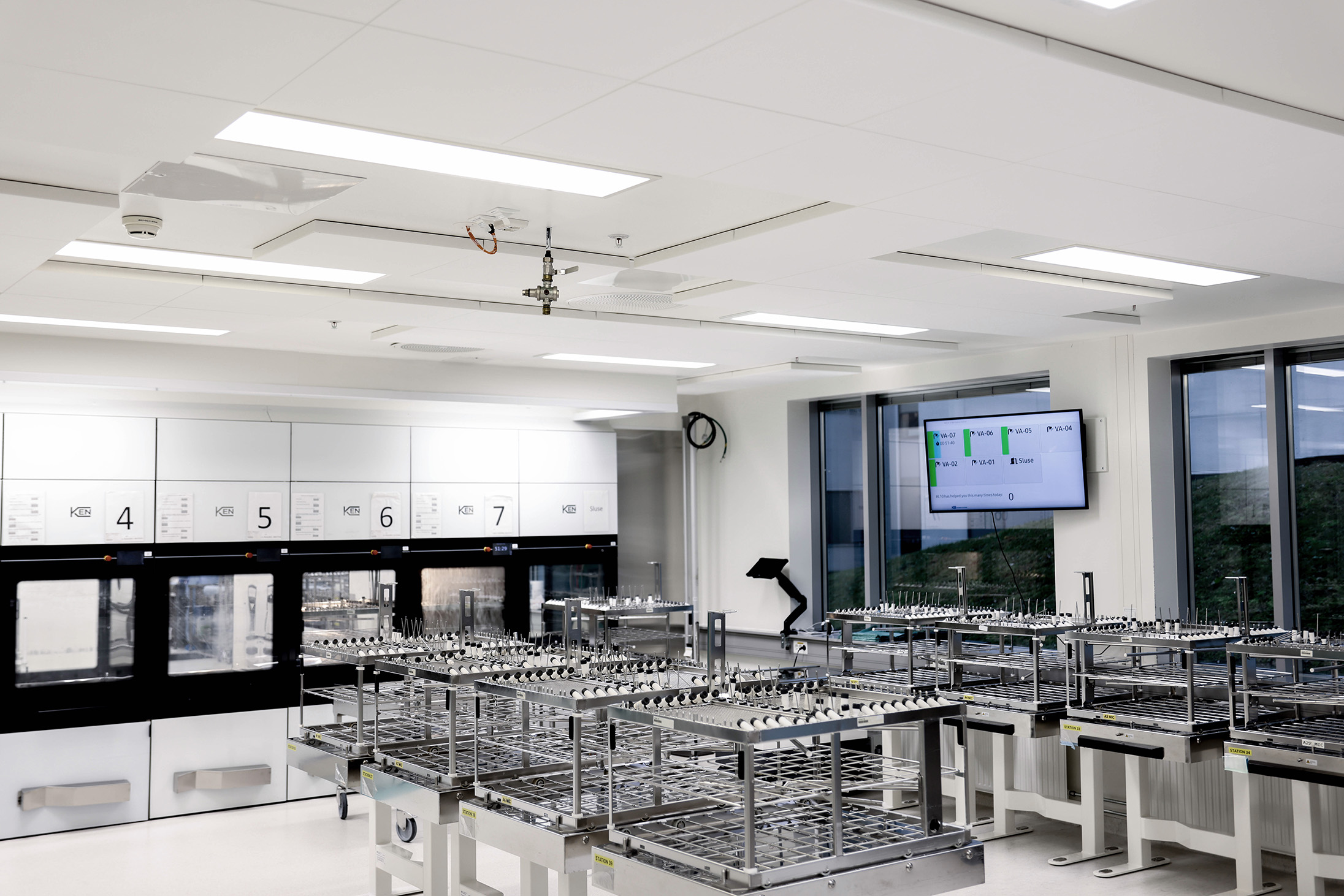 A modern, bright laboratory with multiple empty metal racks, large windows, a wall-mounted monitor displaying data, and numbered equipment units along the back wall. The room appears clean and organised.