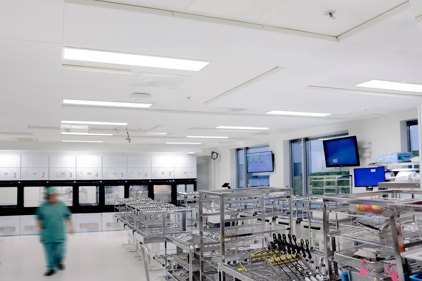 A modern, sterile hospital sterilisation room with metal racks holding surgical instruments. A person in green scrubs walks through the bright, clean space. Monitors and cupboards line the white walls.