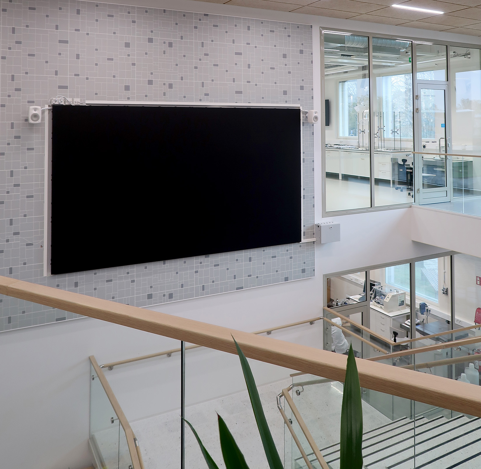 Um grande ecrã preto está montado numa parede revestida a azulejos, acima de uma escadaria, num edifício moderno com laboratórios de paredes de vidro e luz natural. Folhas de plantas verdes são visíveis em primeiro plano.