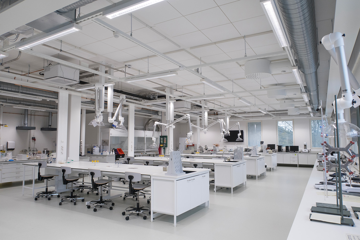 Um laboratório moderno e luminoso, com bancadas brancas, bancos altos, iluminação suspensa e diversos equipamentos científicos. A sala é limpa, espaçosa e maioritariamente vazia, com grandes janelas que deixam entrar luz natural.
