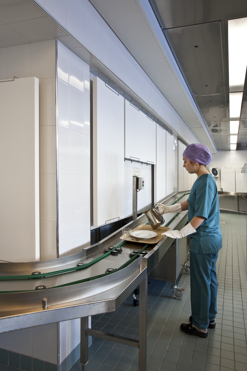 A person in green scrubs and a hair net places a meal tray onto a conveyor belt in a clean, modern hospital or cafeteria kitchen.