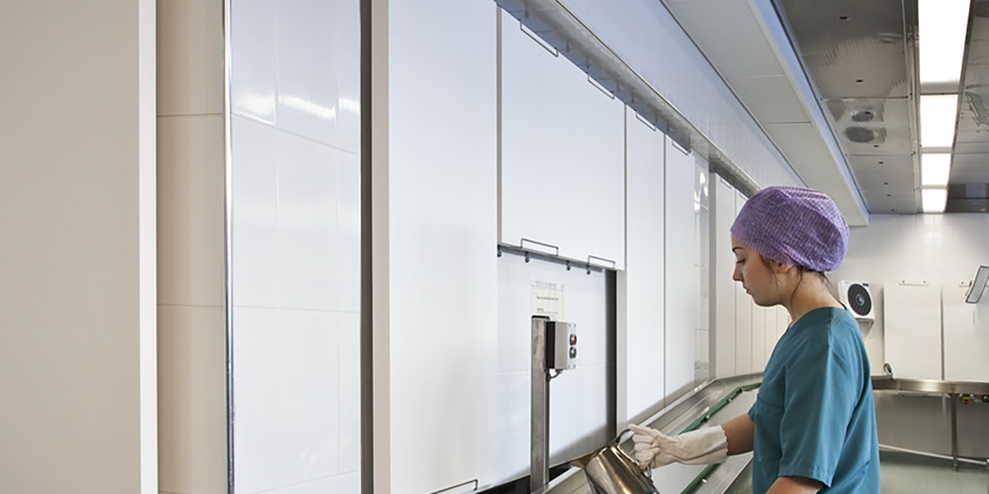 Osoba ubrana w fioletową nakrycie głowy, zielony uniform i białe rękawiczki obsługuje sprzęt w czystej, nowoczesnej kuchni szpitalnej lub stołówce.