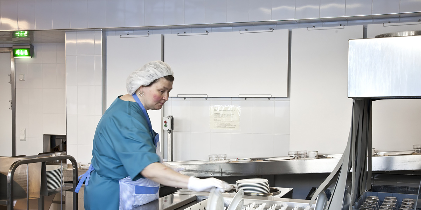 Osoba ubrana w niebieski mundur, siatkę na włosy i rękawiczki wkłada naczynia do zmywarki przemysłowej w komercyjnej kuchni o białych kafelkowych ścianach i metalowych powierzchniach.