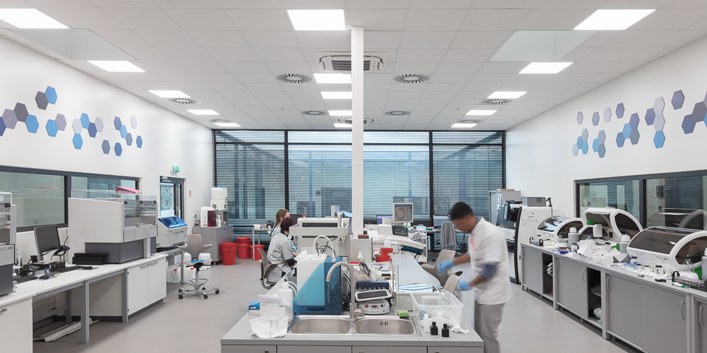 Un laboratorio moderno e luminoso, con scienziati che lavorano ai banchi. Le attrezzature e i computer sono disposti in modo ordinato, e motivi esagonali blu decorano le pareti bianche. Le ampie finestre lasciano entrare la luce naturale.