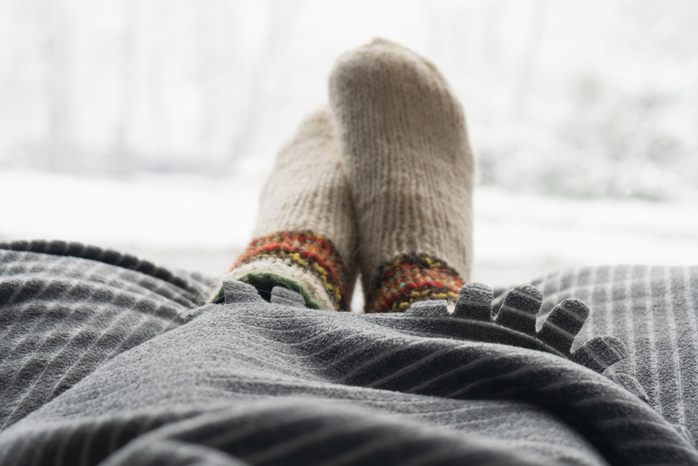 A person with woollen socks and a striped blanket relaxes indoors, with a snowy and blurred winter landscape visible through the window in the background.