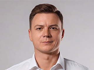 A man with short brown hair, wearing a white dress shirt, stands against a plain light grey background and looks directly at the camera with a neutral expression.