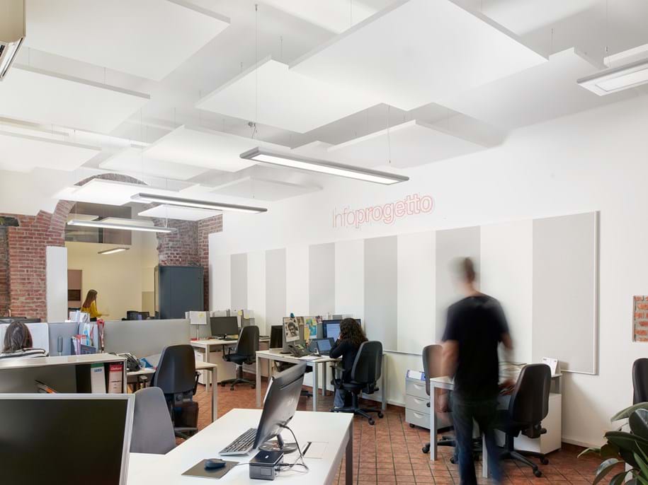 Modern office space with exposed brick walls and ceiling panels. Desks with computers are arranged throughout. A blurred figure walks by, and a person is seated at a desk. The word infoprogetto is visible on the wall.