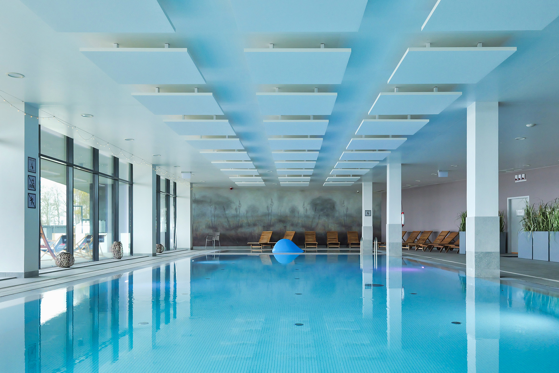 A modern indoor swimming pool with clear blue water, large windows on one side, lounge chairs, potted plants, and white ceiling panels reflecting in the water, creating a calm and inviting atmosphere.