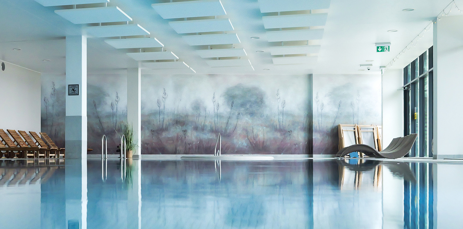 A serene indoor swimming pool with calm water, wooden loungers along the left wall, large windows on the right, and a misty landscape mural on the back wall.